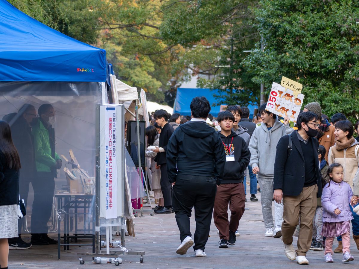 青や白のテントの模擬店が並ぶ並木道を、家族連れや学生が歩いている様子。たこ焼きの手持ち看板が見える