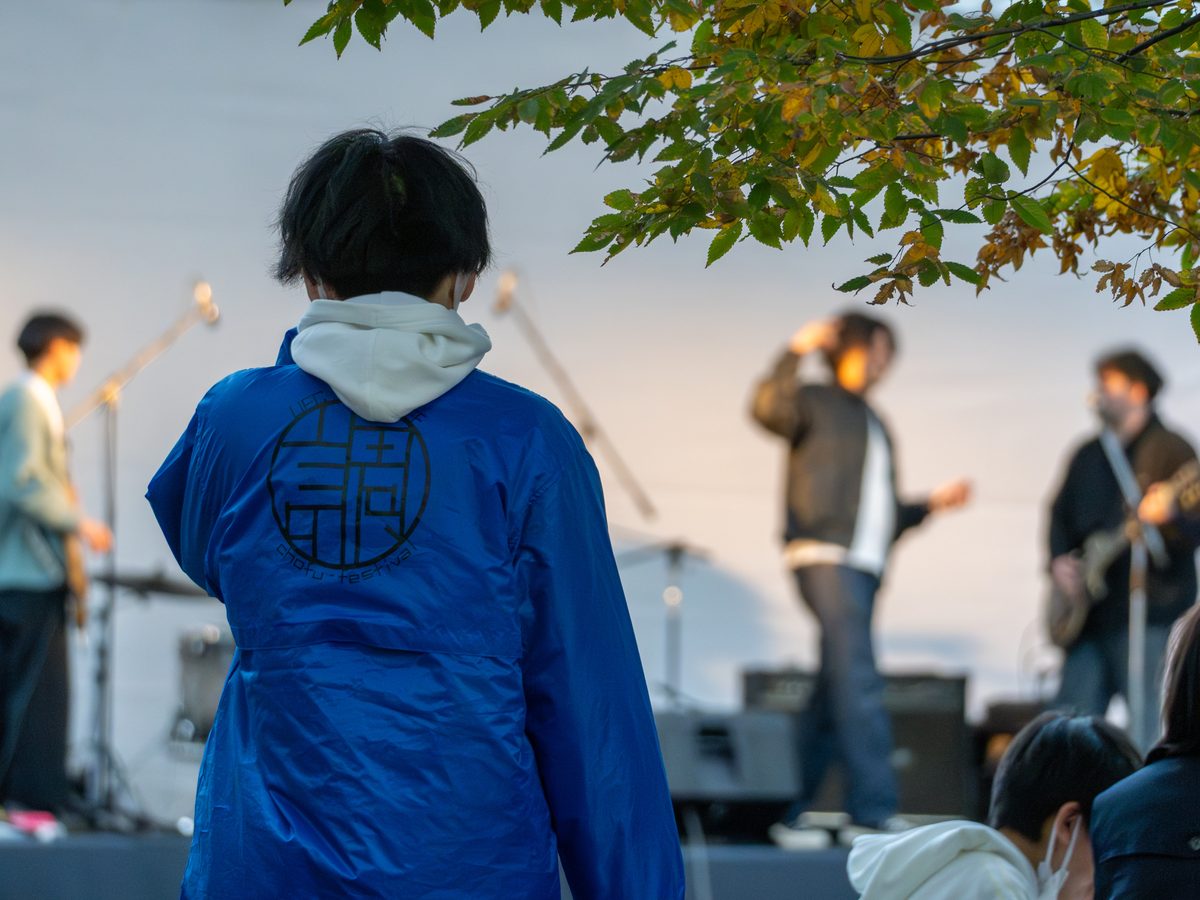 紅葉した木の下の屋外ステージでバンドが演奏し、背中に「調」の字が入った青いはっぴの実行委員が客席側に立っている様子