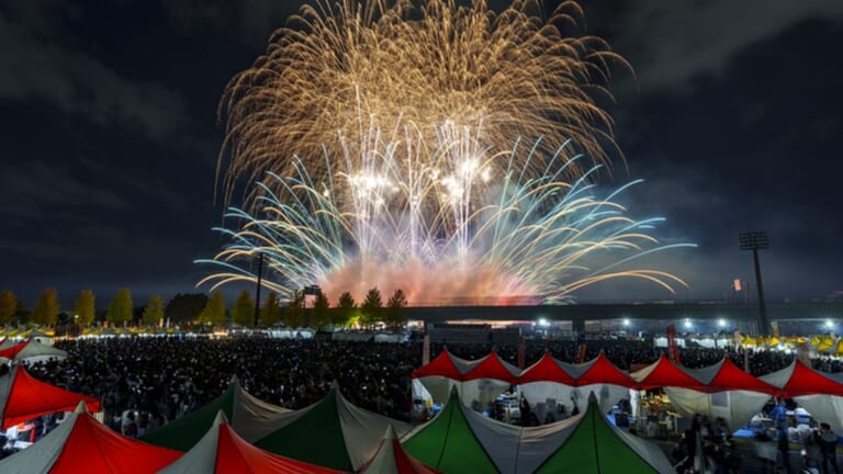 えびな市民まつり2025の花火。海老名運動公園の陸上競技場の上空に金色と白の花火が開き、手前に出店のテントと観客が並ぶ