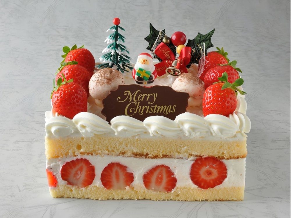 断面に苺が並ぶ四角いショートケーキの上に、苺とサンタ、もみの木、Merry Christmasのプレートを飾った写真