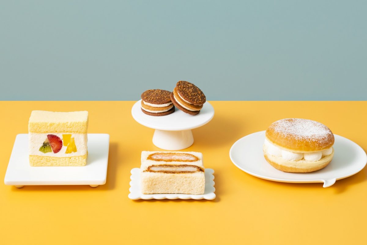 黄色い台の上に、フルーツのケーキサンド、クリームを挟んだクッキー、カステラのサンド、粉糖をかけたドーナツが並ぶ写真