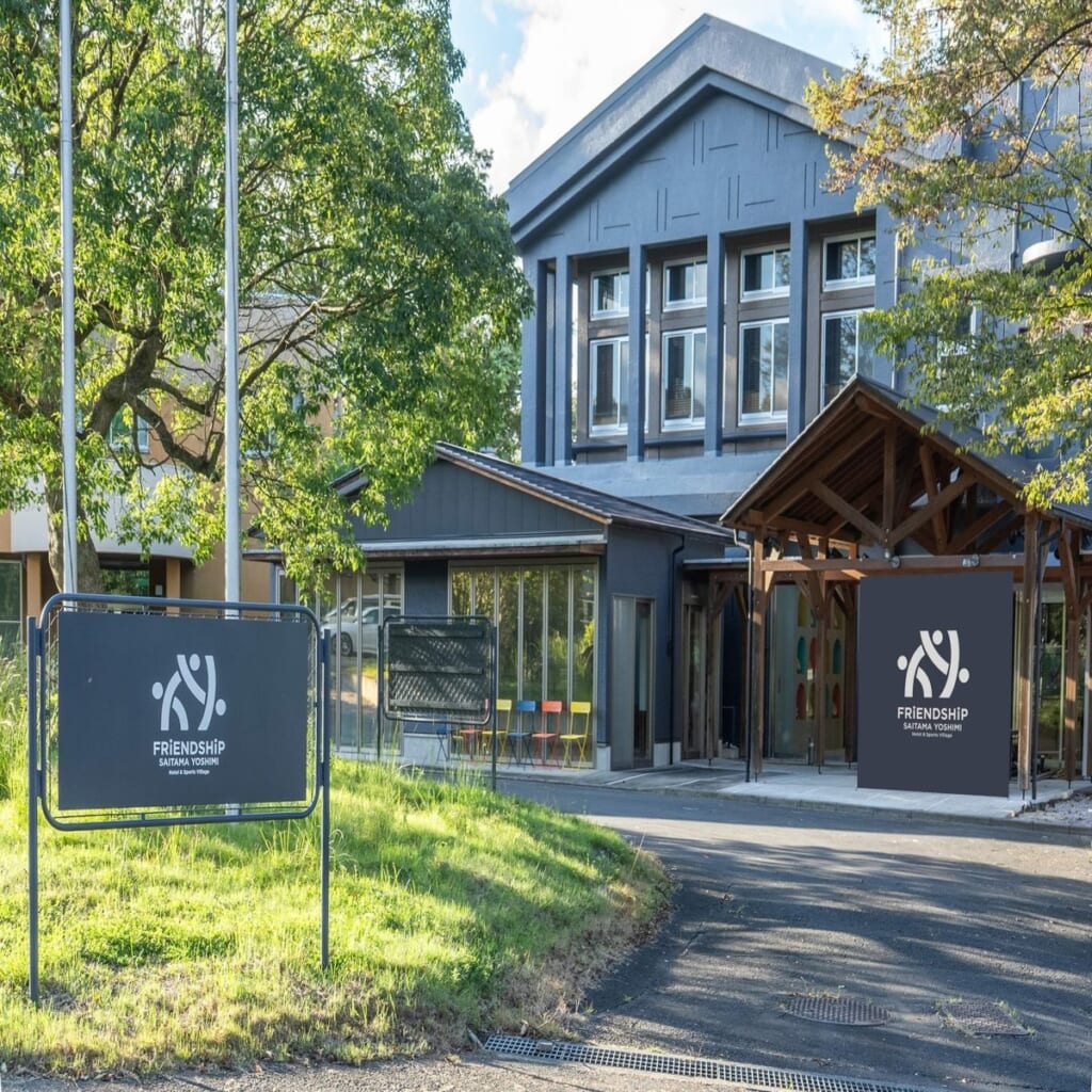 木々に囲まれた濃いグレーの建物と、FRiENDSHiP SAITAMA YOSHIMIのロゴが入った看板が立つ入口