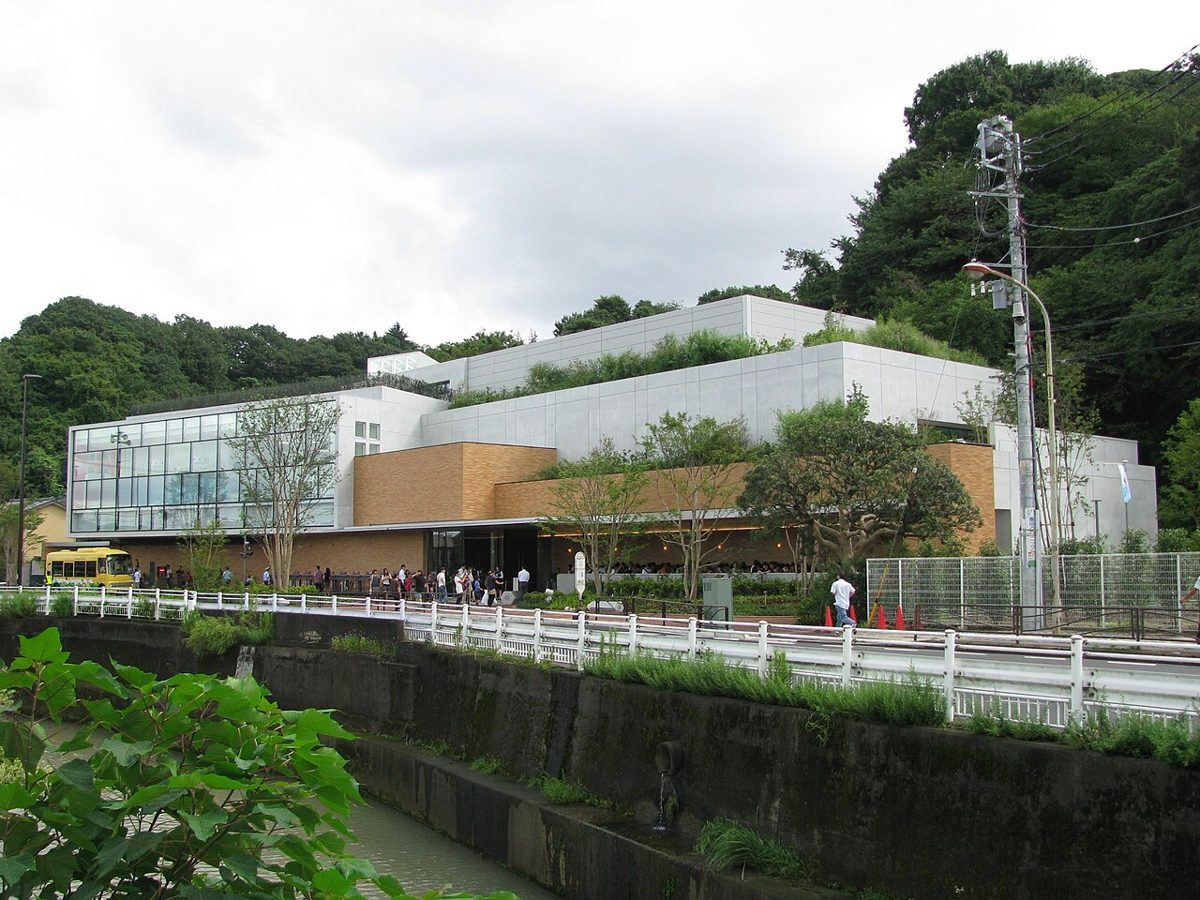 緑の丘を背にした白とガラス張りの建物と、手前の川沿いの柵