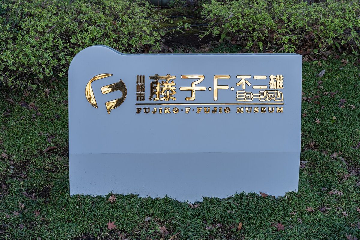 植え込みの前に置かれた白い板に金色の文字で「川崎市 藤子・F・不二雄ミュージアム」