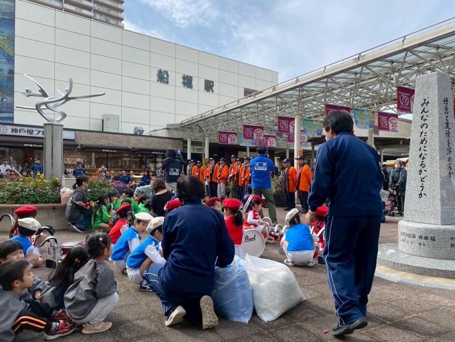 船堀駅前の広場に座る保育園児たちと、オレンジ色の服を着た参加者が並ぶパレード出発式