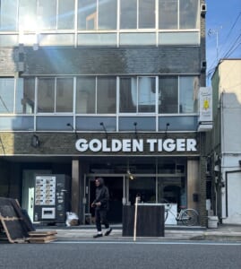レンガ調のビルの1階に「GOLDEN TIGER」の白い文字看板が付いた新店舗の入口。左側に冷凍麺の自動販売機がある