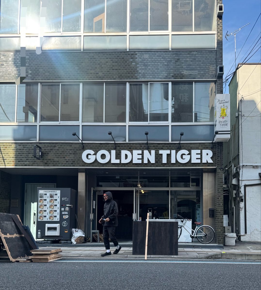 レンガ調のビルの1階に「GOLDEN TIGER」の白い文字看板が付いた新店舗の入口。左側に冷凍麺の自動販売機がある