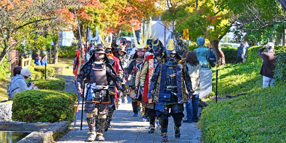 色づいた木々の下の石畳を、兜と鎧を着けた武者たちが列になって歩く様子