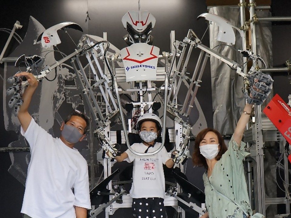 銀色の大型ロボットの前で、操縦用の装置を着けた子どもと大人2人が手を挙げている写真