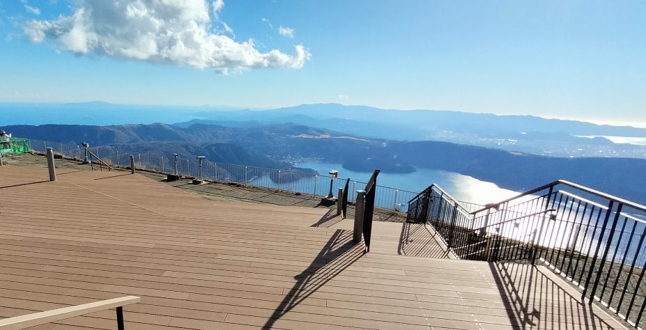 晴れた日中の山頂展望広場のウッドデッキと手すり。奥に芦ノ湖と外輪山が見える