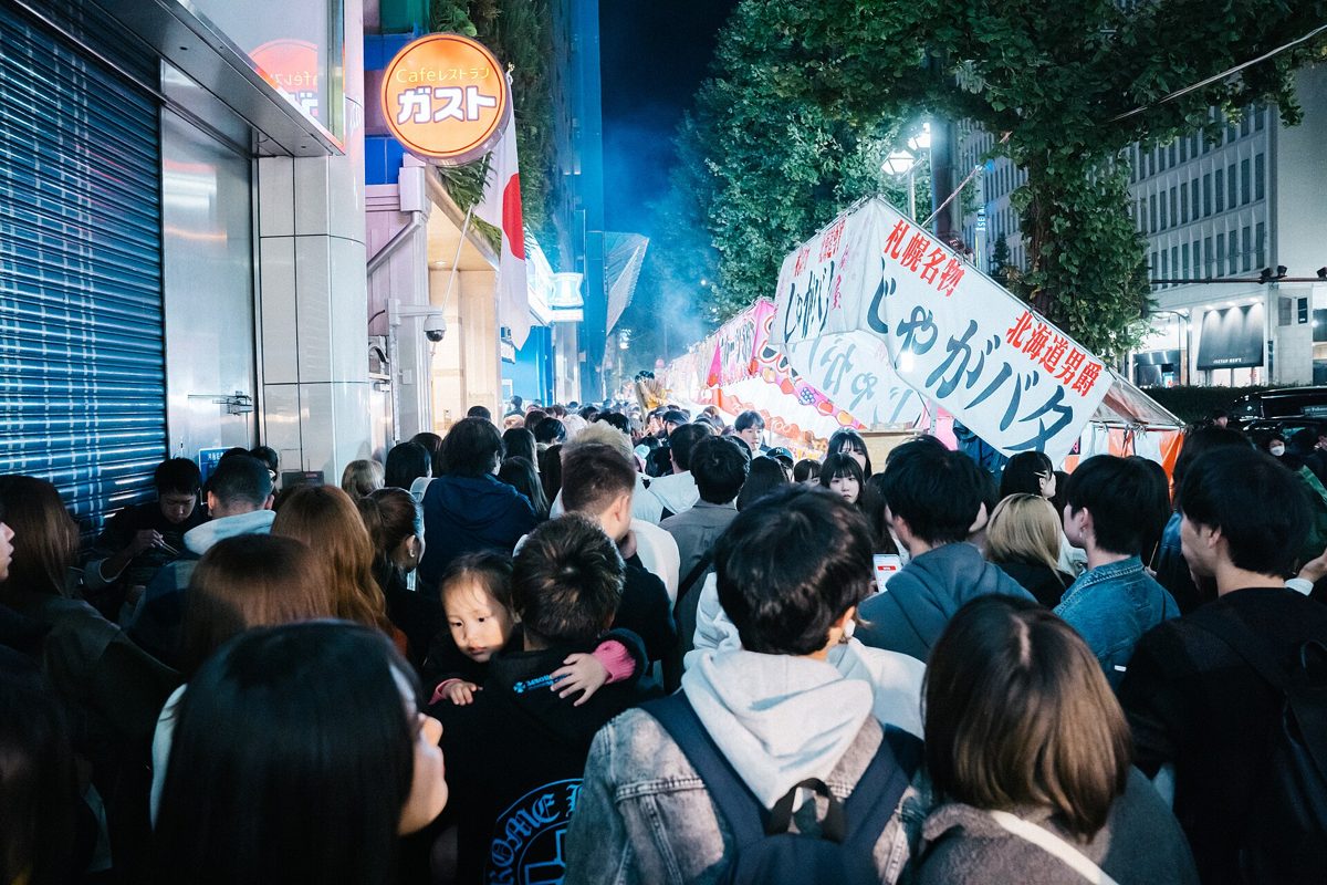 夜の新宿の歩道を、露店の看板の下で大勢の人が肩を寄せ合って進んでいる