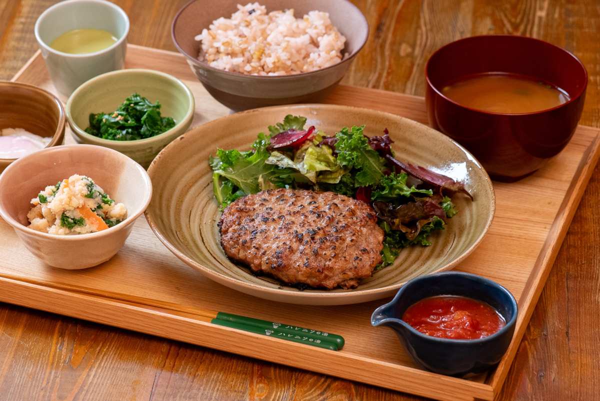 木のお盆に、サラダを添えたハンバーグの皿、赤いソース、ごはん、味噌汁、小鉢が並ぶ