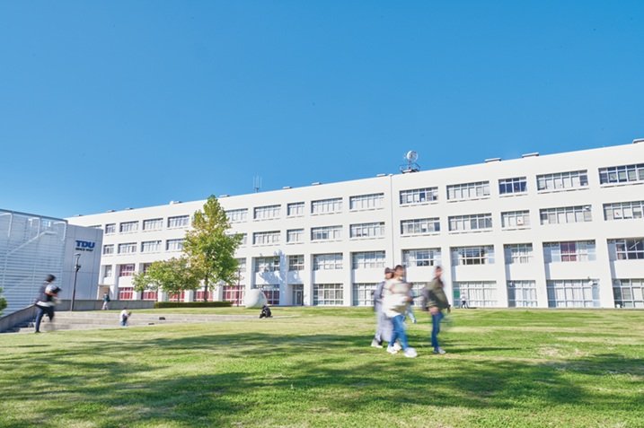 青空の下、芝生の広場の奥に白い4階建ての校舎が横に長く続き、手前を学生が歩くキャンパスの写真