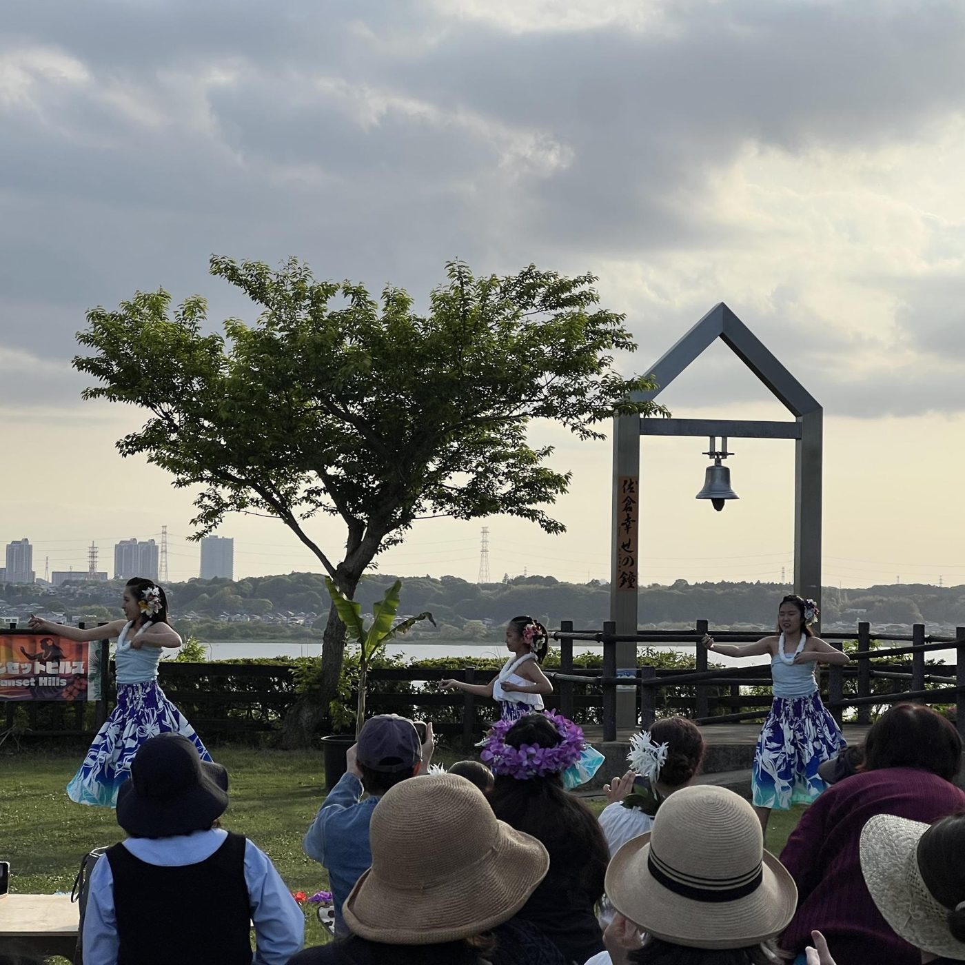 印旛沼サンセットヒルズの芝生で、青いスカートの衣装でフラを踊る女性たちと、帽子をかぶって見る観客。背後に鐘のついたモニュメントと印旛沼