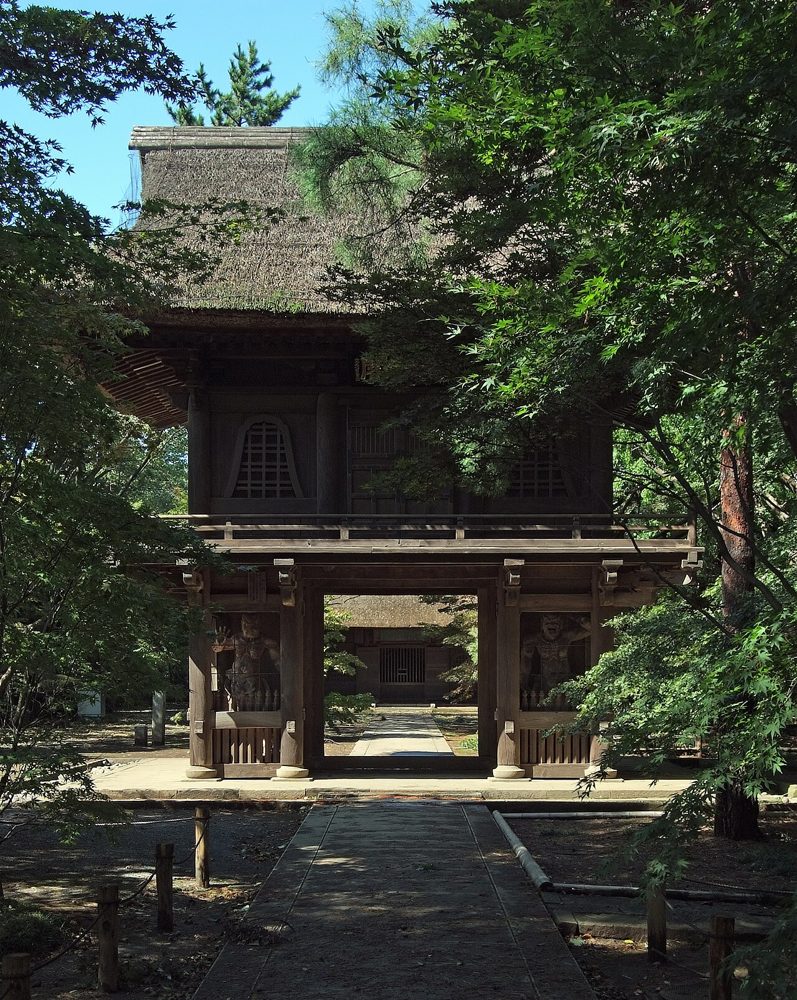 木々に囲まれた参道の先に建つ、茅葺屋根の二層の山門の写真