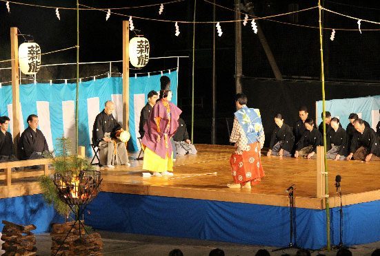 過去のヘリテイジ薪能の様子。屋外の特設舞台で装束姿の演者2人が向き合い、舞台脇にかがり火と「薪能」の提灯