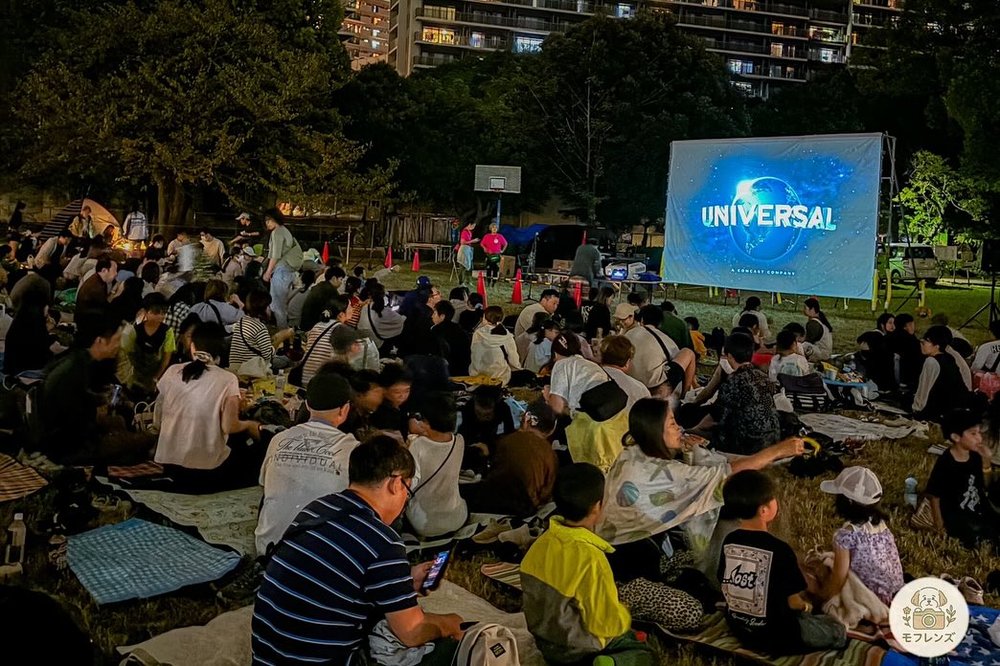 2025年の第4回ひがしこうえんマルシェ星空映画祭。夜の稲毛東公園の芝生にシートを敷いて座った大勢の来場者と、大型スクリーン