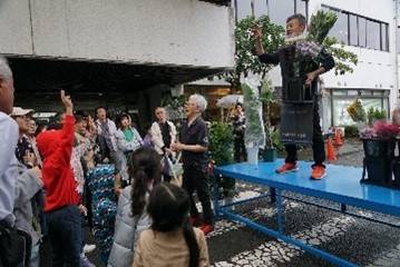 屋外の台の上で花束を掲げるせり人と、手を挙げて参加する来場者