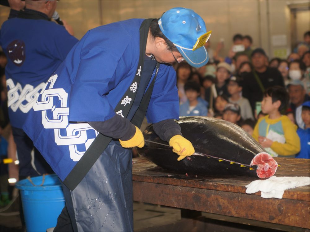 青い法被の職人が台の上のマグロに包丁を入れ、子どもを含む見物客が囲んでいる