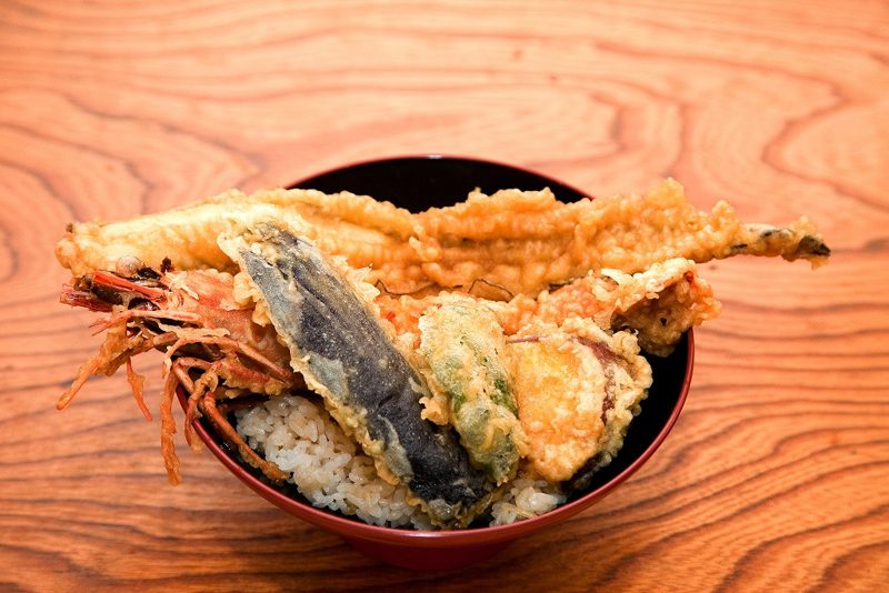 木のテーブルに置かれた丼に、大きな海老と穴子、野菜の天ぷらがのっている