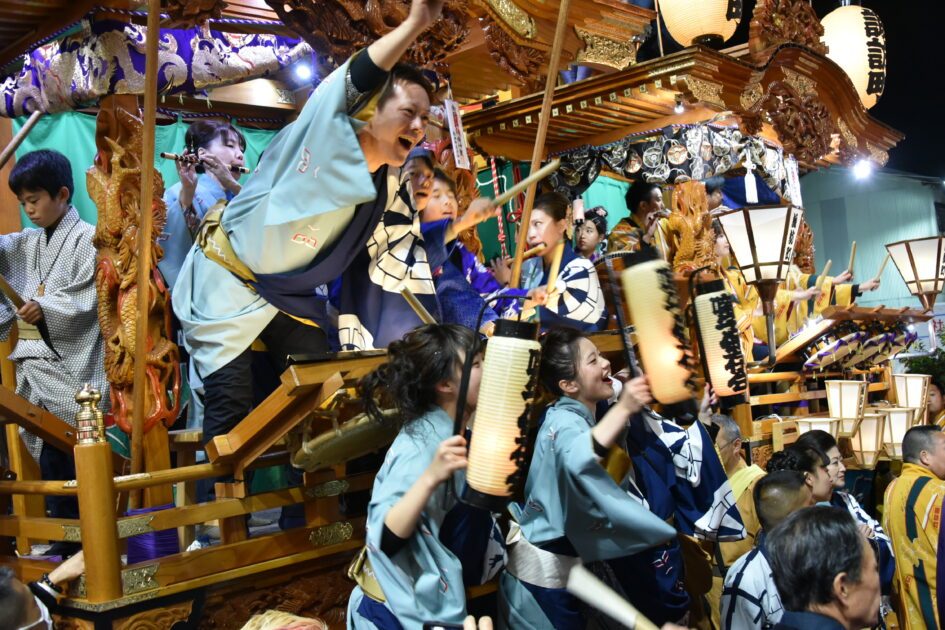 夜、彫刻で飾られた山車の上で囃子方が笛や太鼓を鳴らし、手前で揃いの浴衣を着た女性たちが提灯を掲げて声を上げている写真