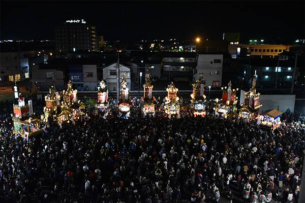 夜、明かりのともった山車が横一列に並び、その前の広場を大勢の見物客が埋めている写真