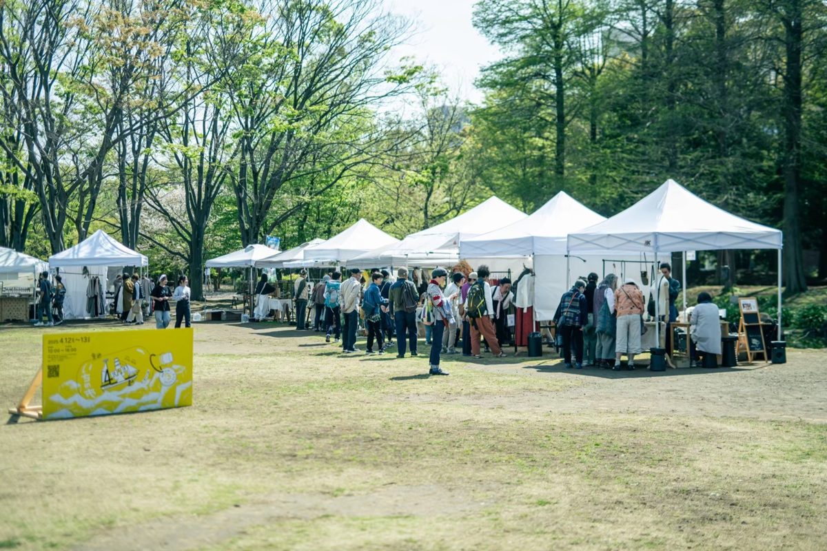 公園の芝生に白いテントのブースが並び、来場者が作品を見て回る様子