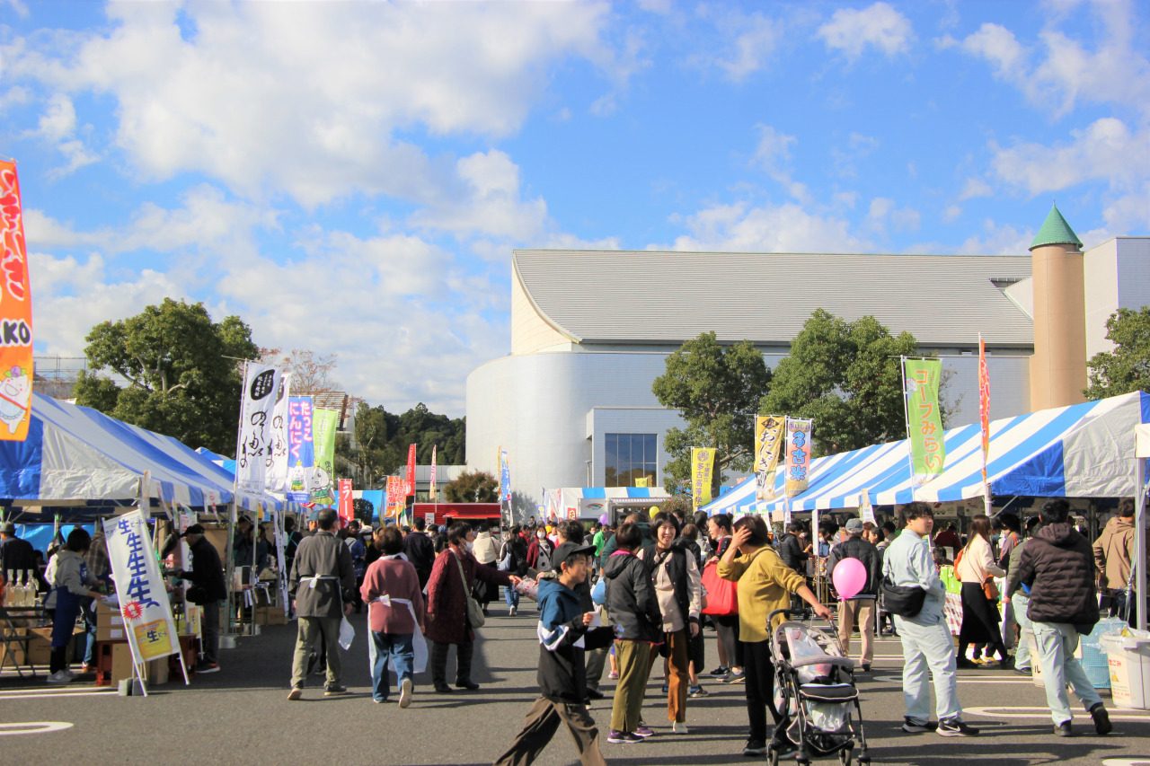 青空の下、多古町コミュニティセンターの白い建物の前に青と白のテントとのぼり旗が並び、ベビーカーを押す人や家族連れが歩くいきいきフェスタTAKOの会場