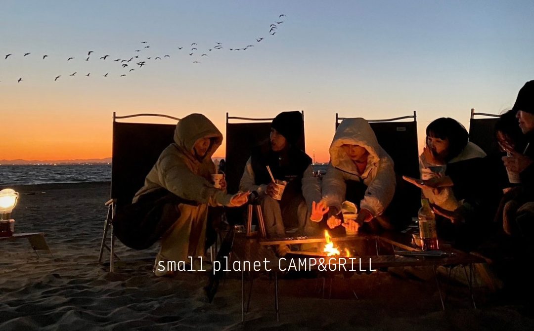 夕焼けの海を背に、砂浜で椅子に座った人たちが焚き火台を囲んで手をかざし、空に鳥の群れが飛んでいる写真。下部に「small planet CAMP&GRILL」の文字