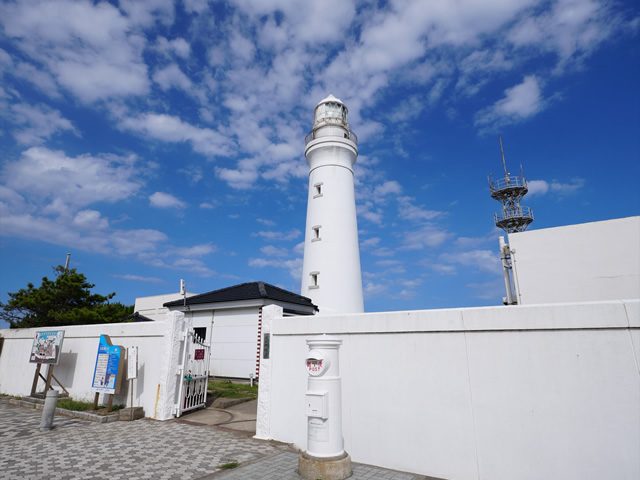 白い塀の門の前に白いポストが立ち、奥に白い灯台