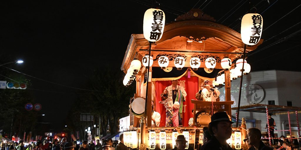 夜の通りで提灯をともした賀美町の山車。屋根の下で囃子の人形が飾られ、周りに見物客が集まっている