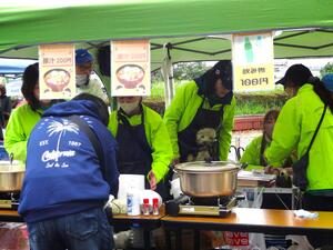 緑のテントの模擬店で、蛍光色のベストを着たスタッフが鍋の豚汁を用意している。豚汁と「飲み物100円」の貼り紙