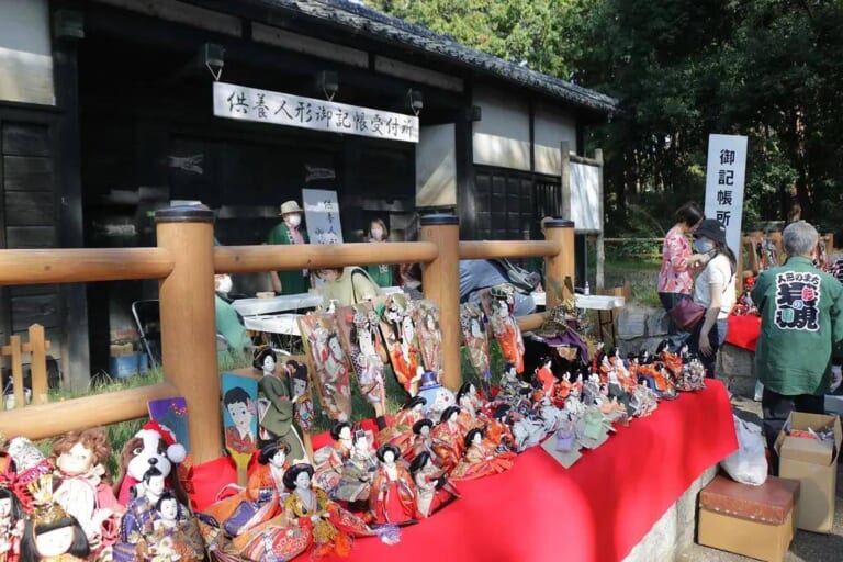 岩槻城址公園の黒門前に並べられた、供養を待つひな人形や羽子板、ぬいぐるみ。奥に「供養人形御記帳受付所」の看板と受付の人たち