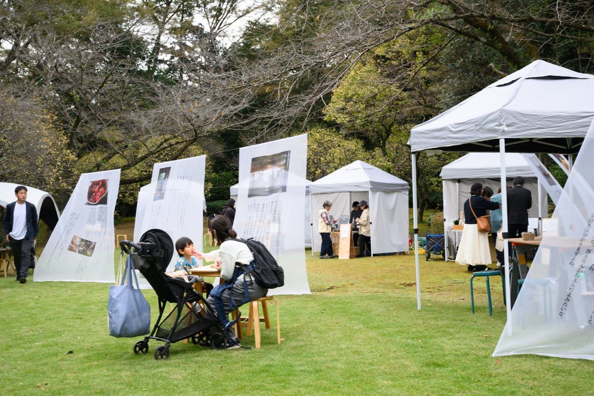 芝生の公園に白いテントが並び、ベビーカーの家族がテーブルで休んでいる