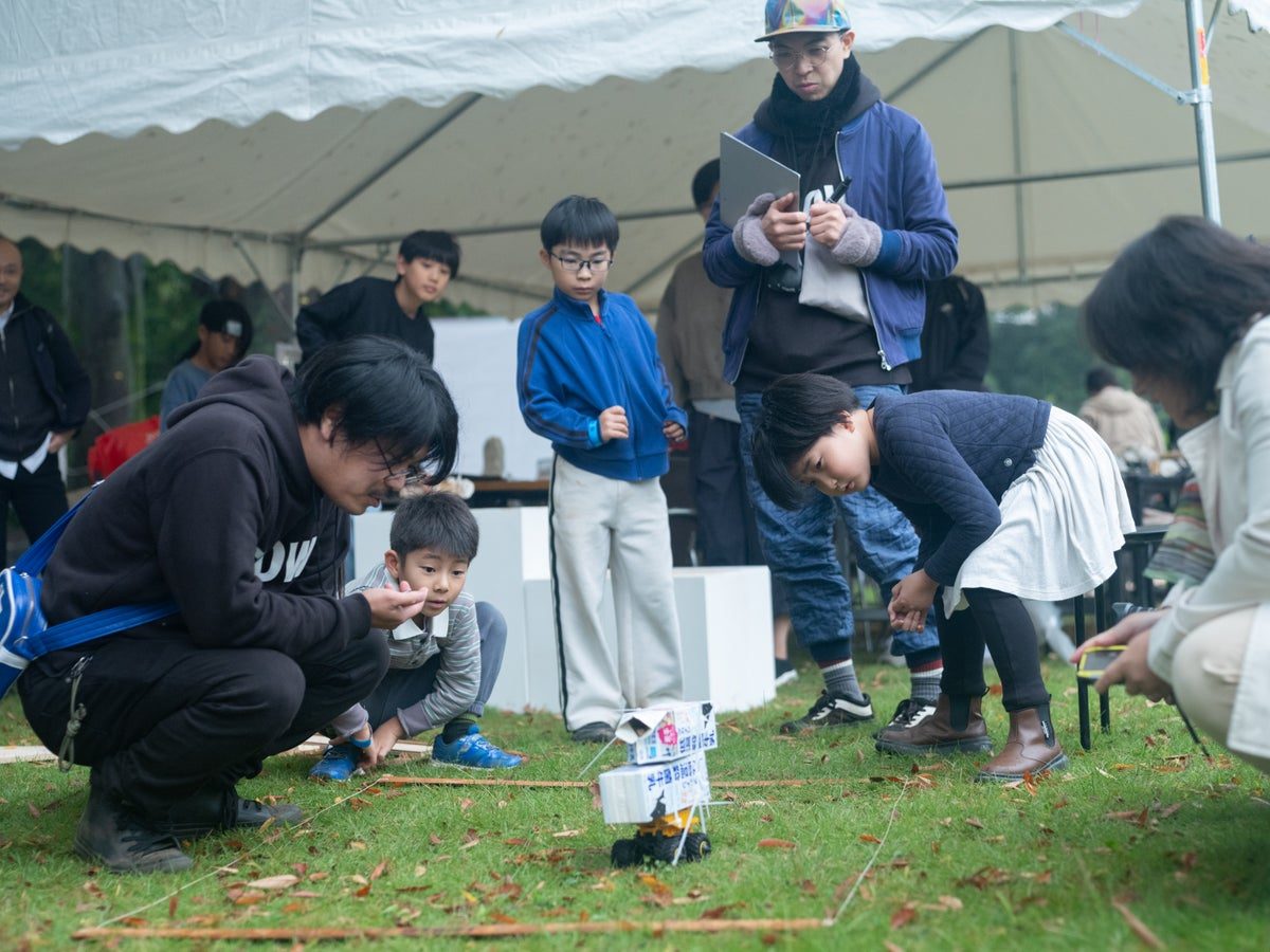 芝生の上にしゃがんだ子どもと大人が、手作りのミニ四駆の走りを見守っている