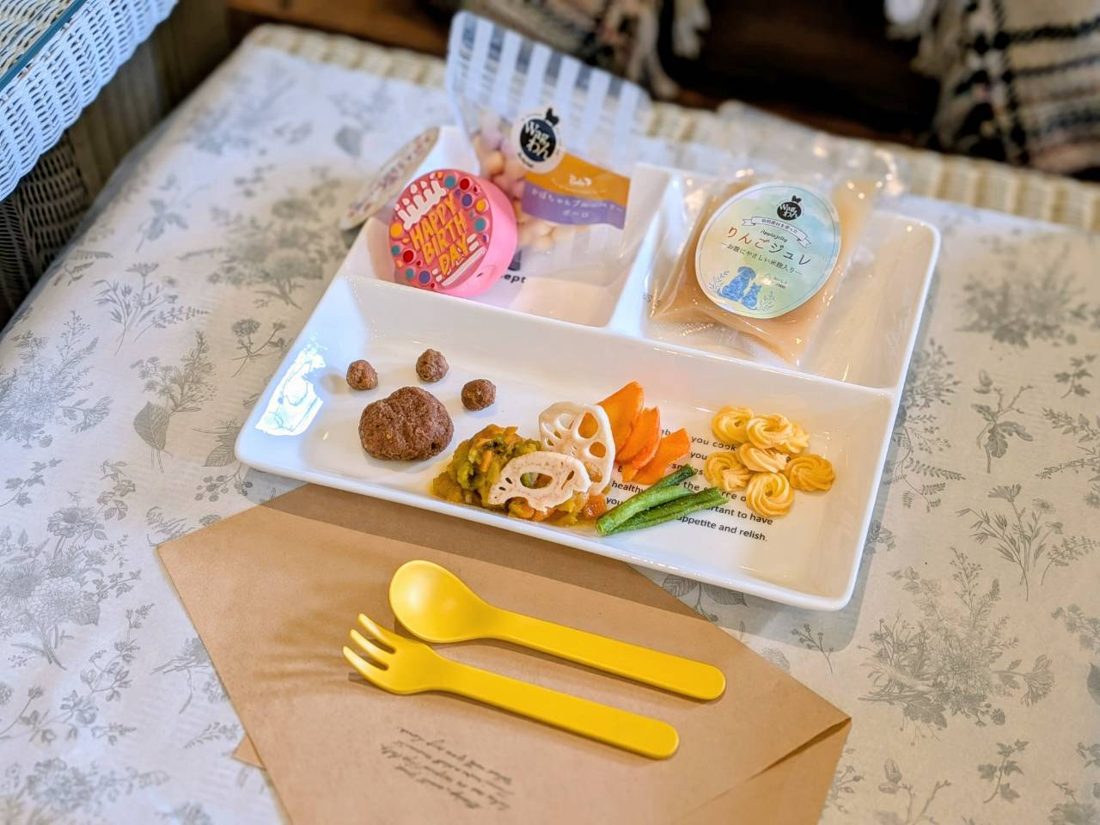 白い仕切り皿に野菜や肉、ボーロのような粒が盛られ、袋入りのおやつが添えられた犬用のプレートと、黄色いフォークとスプーン