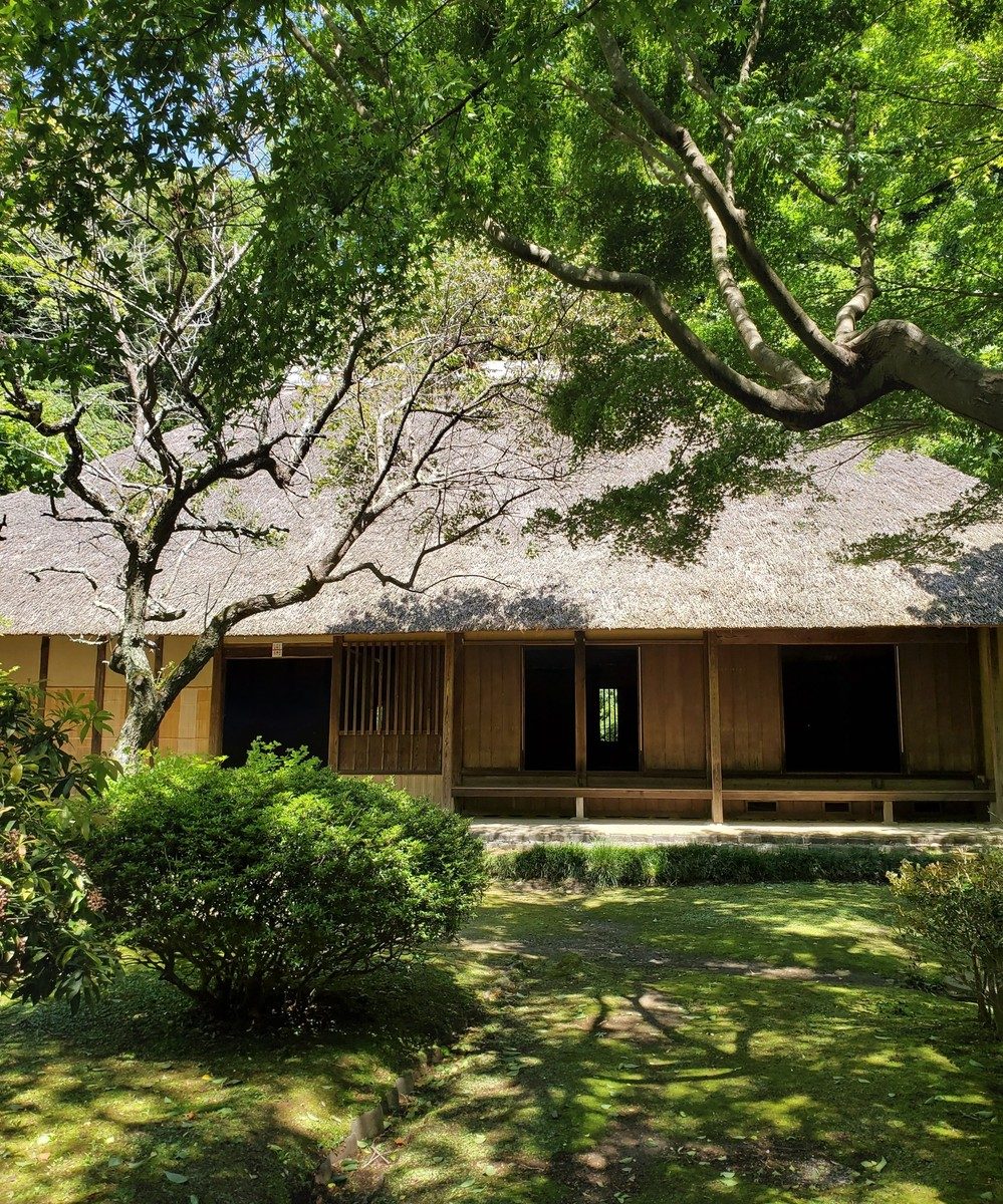 新緑の木々の下にある、茅葺屋根の大きな古民家の正面
