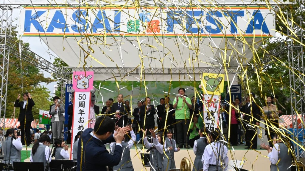 第50回葛西まつりの開会式。KASAI FESTAと書かれたステージで金色のテープが舞い、手前で吹奏楽部が演奏している