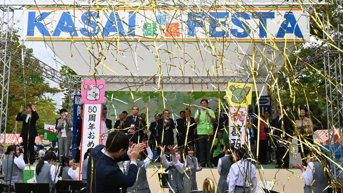 第50回葛西まつりの開会式。KASAI FESTAと書かれたステージで金色のテープが舞い、手前で吹奏楽部が演奏している