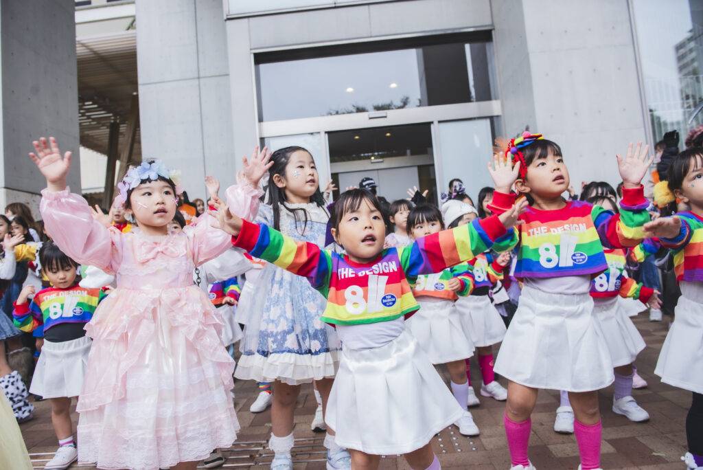 仮装した子どもたちが建物の前の広場で両手を上げて踊っている写真。ピンクのドレスや虹色のそろいの衣装の子どもが並んでいる