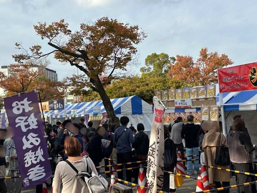 色づいた木の下の大型テントと、その周りで食べたり並んだりする大勢の来場者。地面にはシートと荷物