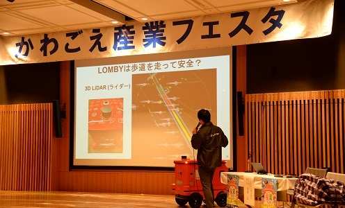「かわごえ産業フェスタ」の横断幕があるステージで、自動走行ロボットLOMBYのスライドを映して講演する登壇者と、赤いロボット