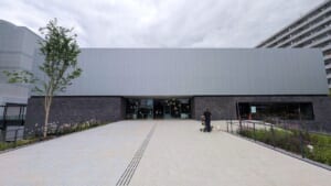 川口市立美術館の外観。灰色の金属の外壁とれんが調の壁の建物へ、植栽に挟まれた広い通路が続いている
