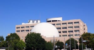 白いプラネタリウムのドームと、屋上に銀色の天文台ドームが3つ並ぶ建物を、青空の下で木々の向こうから撮った写真