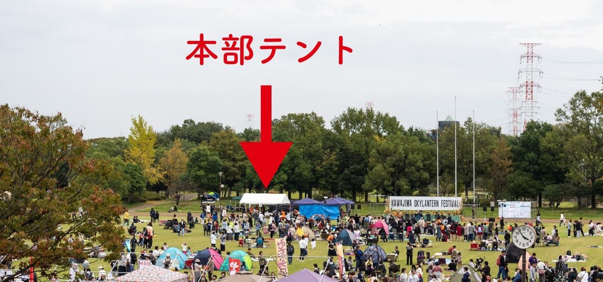 芝生の広場にテントやレジャーシートが広がり、奥に白い本部テントと「KAWAJIMA SKYLANTERN FESTIVAL」の看板を掲げたステージが見える昼間の写真。本部テントを赤い矢印で示している