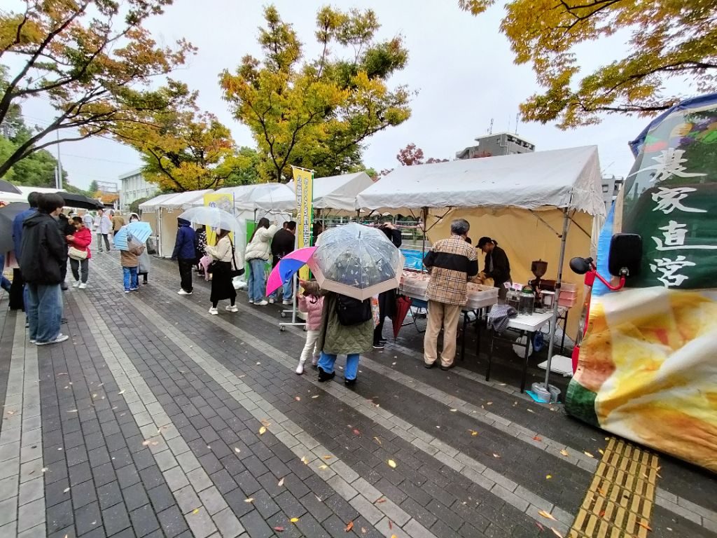 雨の中、紅葉した木の下に白いテントの出展ブースが並び、傘を差した来場者が歩くNEC玉川事業場の公開空地