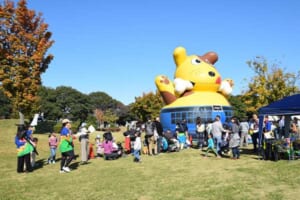 晴れた秋の芝生広場に黄色い犬の大型エア遊具が立ち、子ども連れの来場者やテントが並ぶ川里中央公園の写真