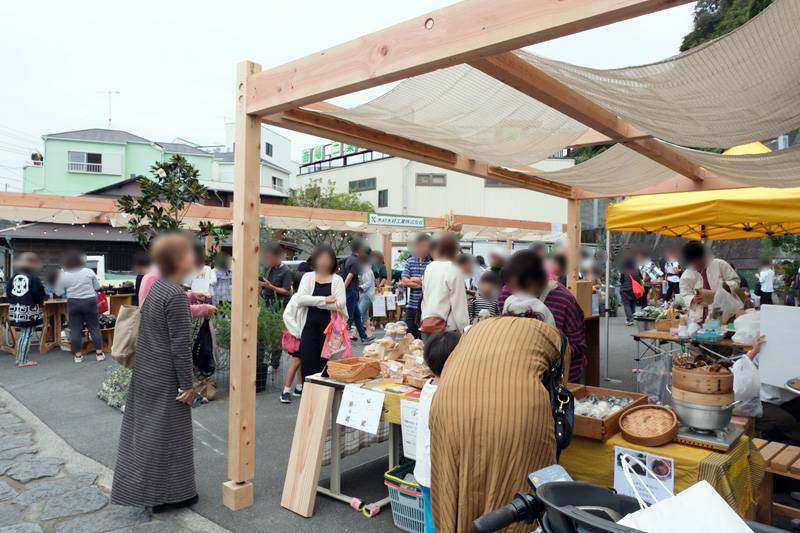 木の柱と布の屋根のブースが並び、来場者が焼き菓子や木工品のテーブルを見ている。人物の顔はぼかし加工されている