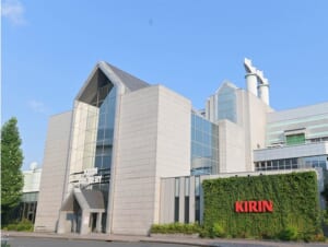 青空の下、白い建物の前に緑の植え込みと赤い「KIRIN」の文字がある工場の入口