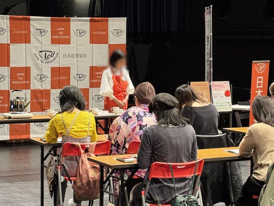 日本紅茶協会のロゴが並ぶパネルの前で、講師の話を聞きながら机でお茶を味わう参加者たち